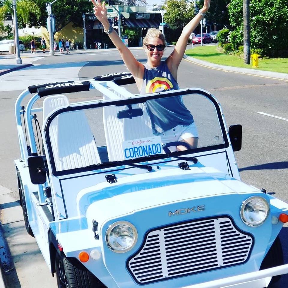 A woman standing in a light blue Moke with her arms raised on a palm-lined street