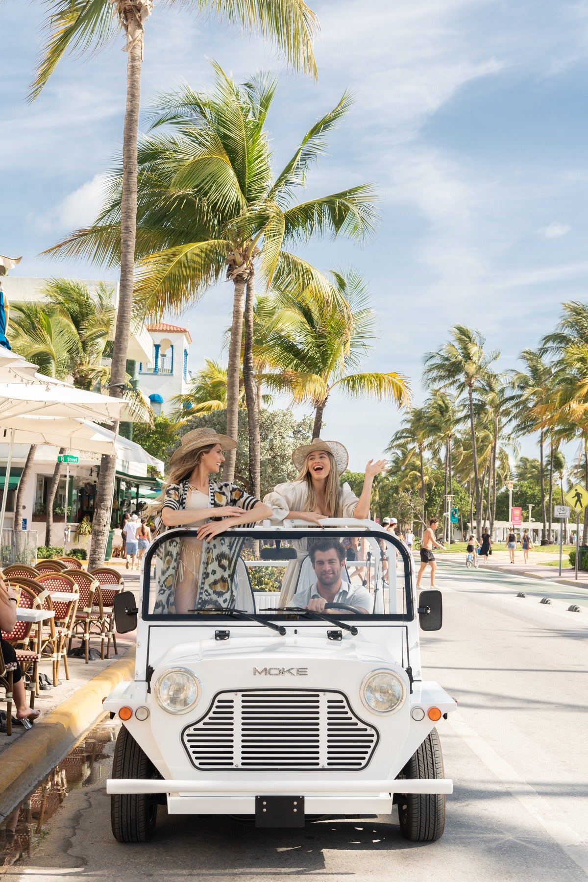 Two people in a white Moke on a palm-lined Miami Beach street
