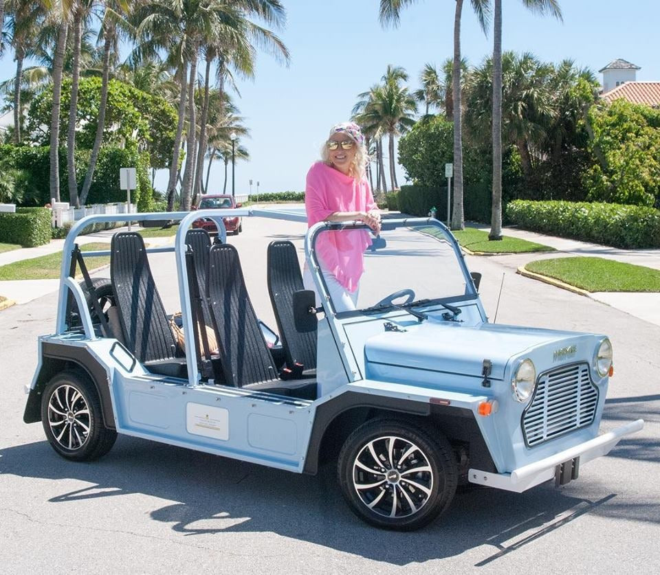 A woman standing beside a light blue Moke on a palm-lined Palm Beach street